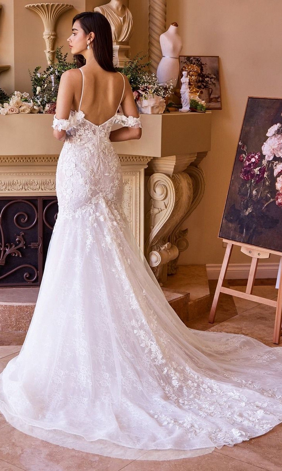 A woman in the Andrea and Leo WL051 long wedding dress, a fitted mermaid gown with lace applique and a long train, stands facing away in an elegant room with floral decor and an easel displaying a painting.